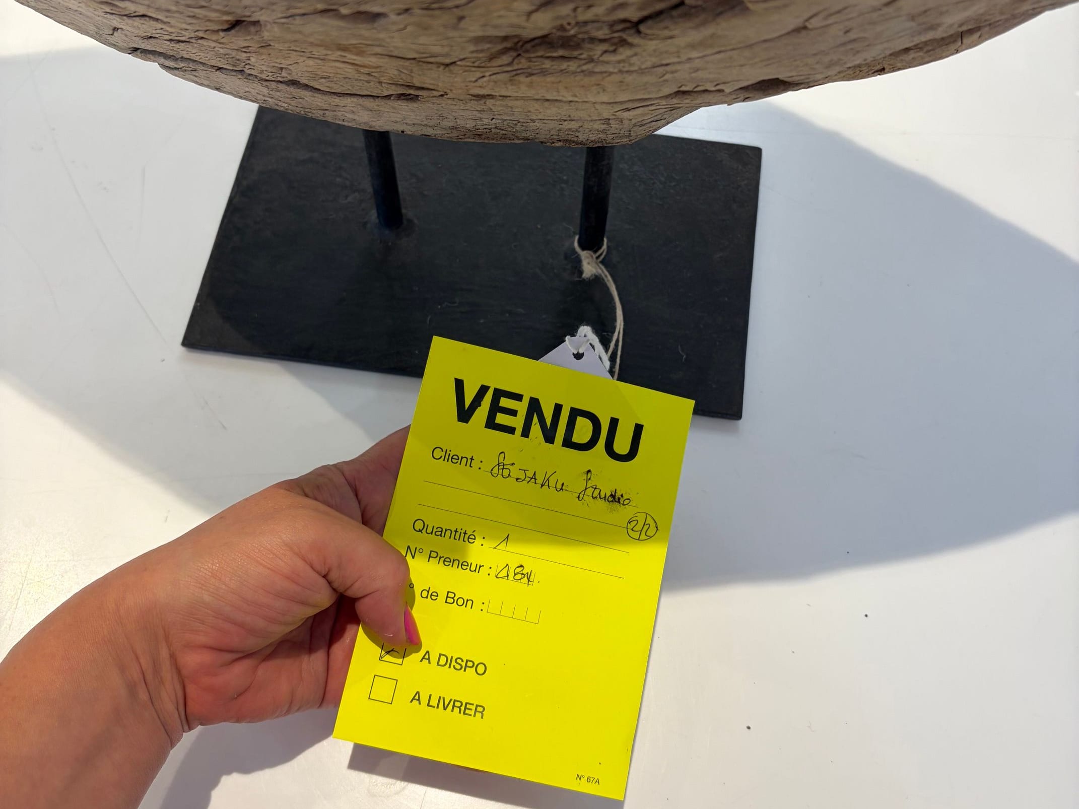 A hand holding a yellow 'Vendu' sold tag marked Seijaku Studio beneath a weathered wood piece on a metal stand