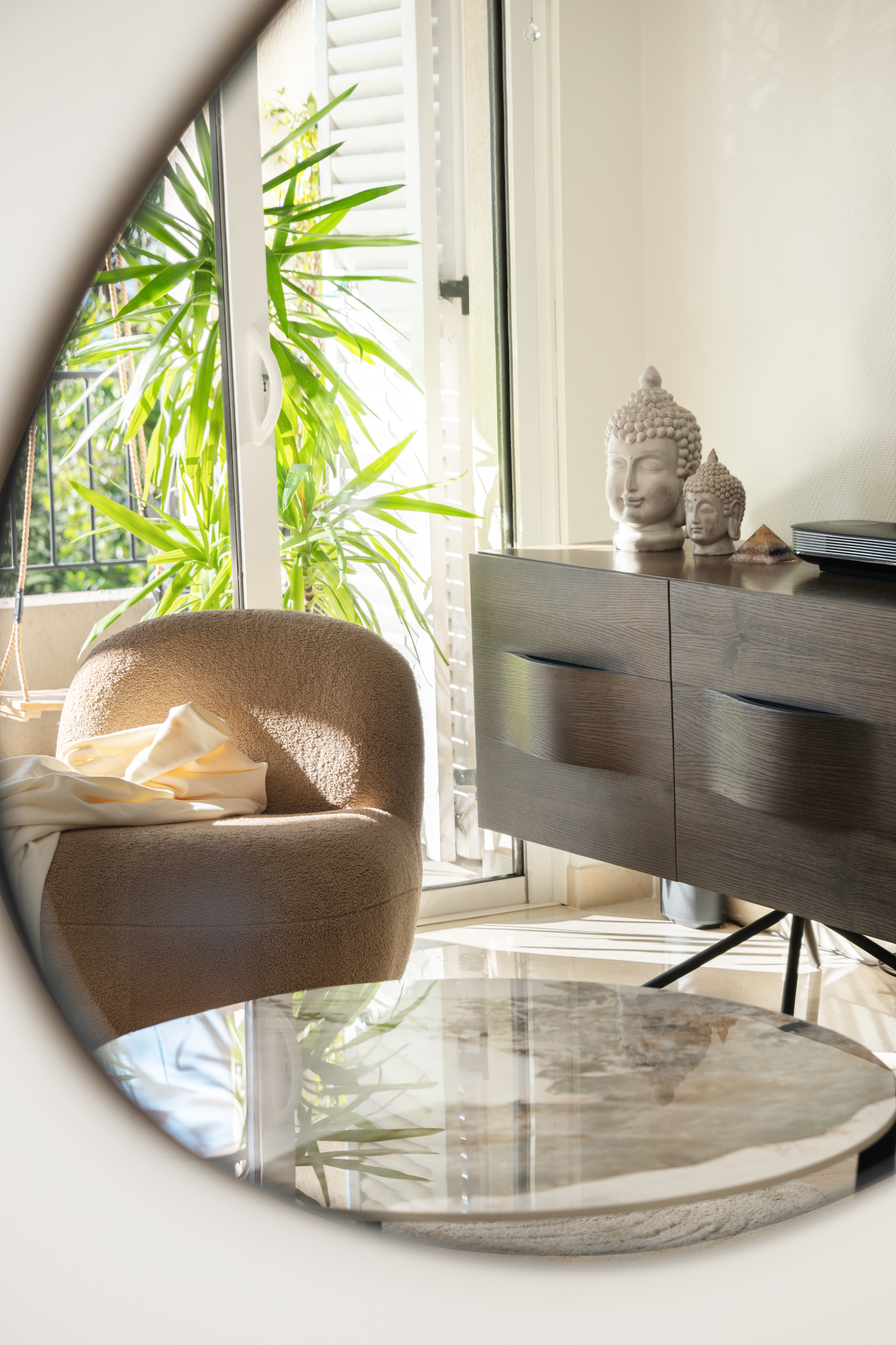 A round mirror reflecting a boucle armchair, a plant, and a sideboard styled with Buddha head sculptures
