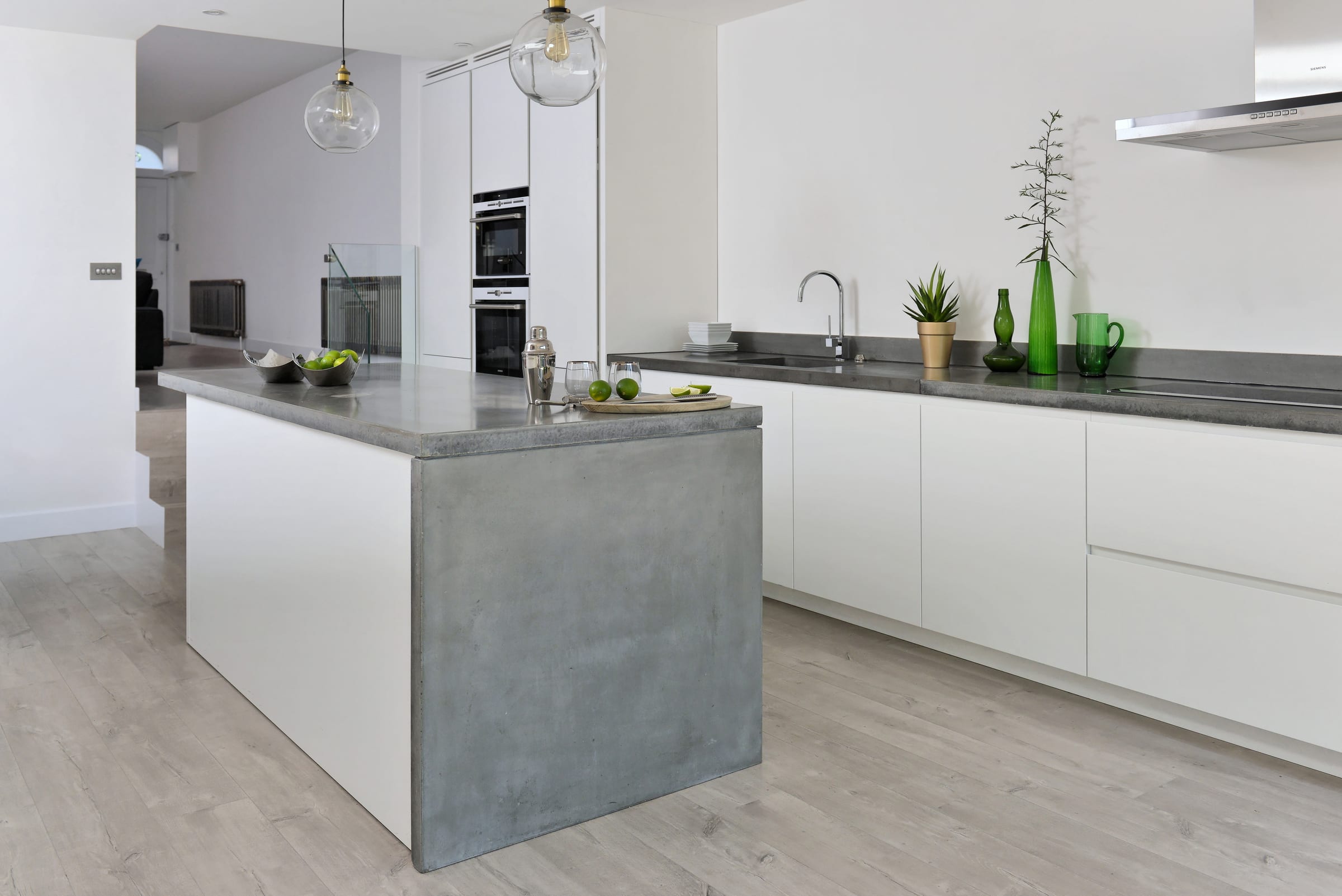 White kitchen with a polished concrete island, glass pendant lights, and green glassware