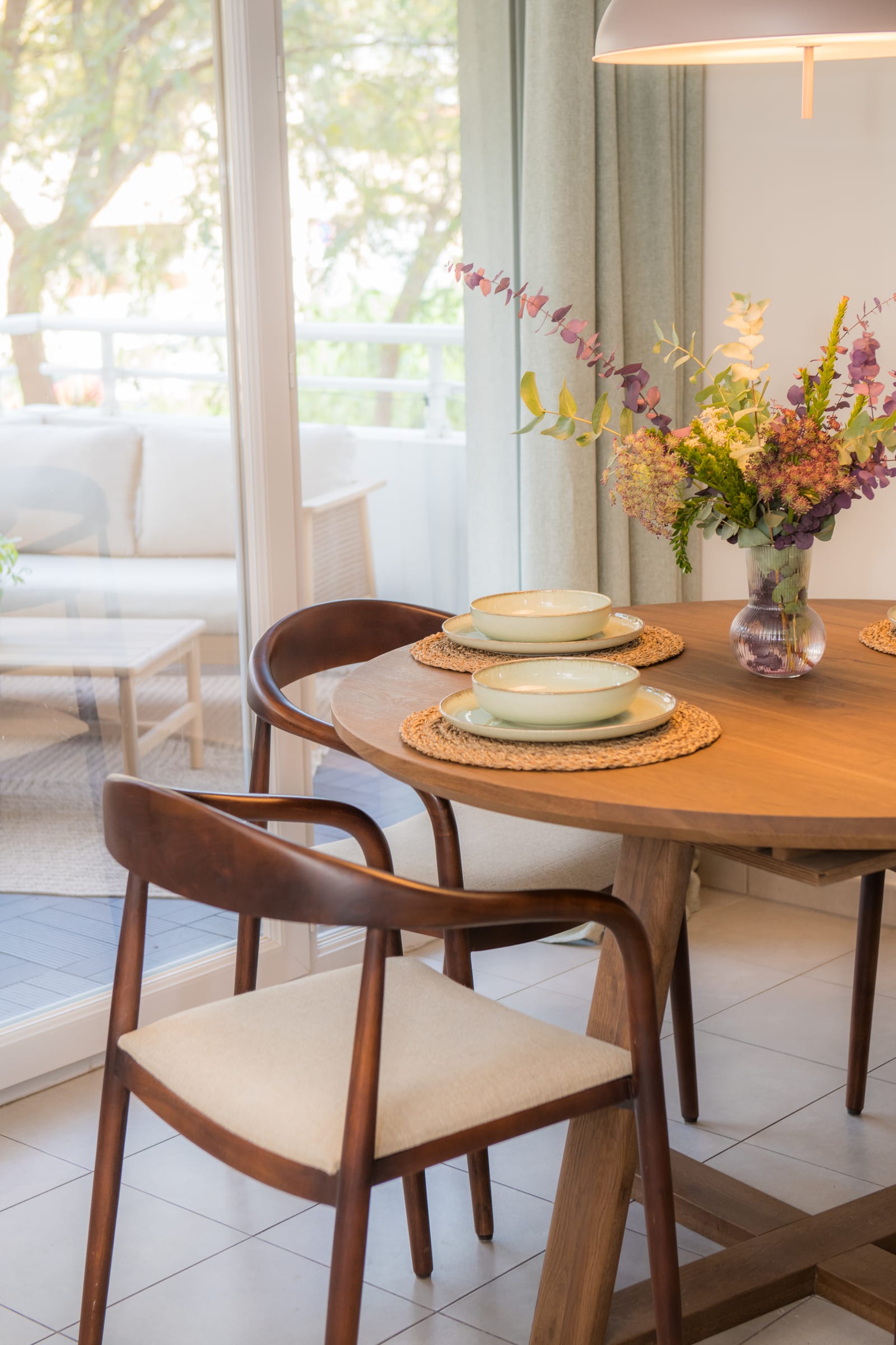 Round walnut dining table set with ceramic bowls, woven placemats, and a dried flower arrangement, with curved wooden armchairs