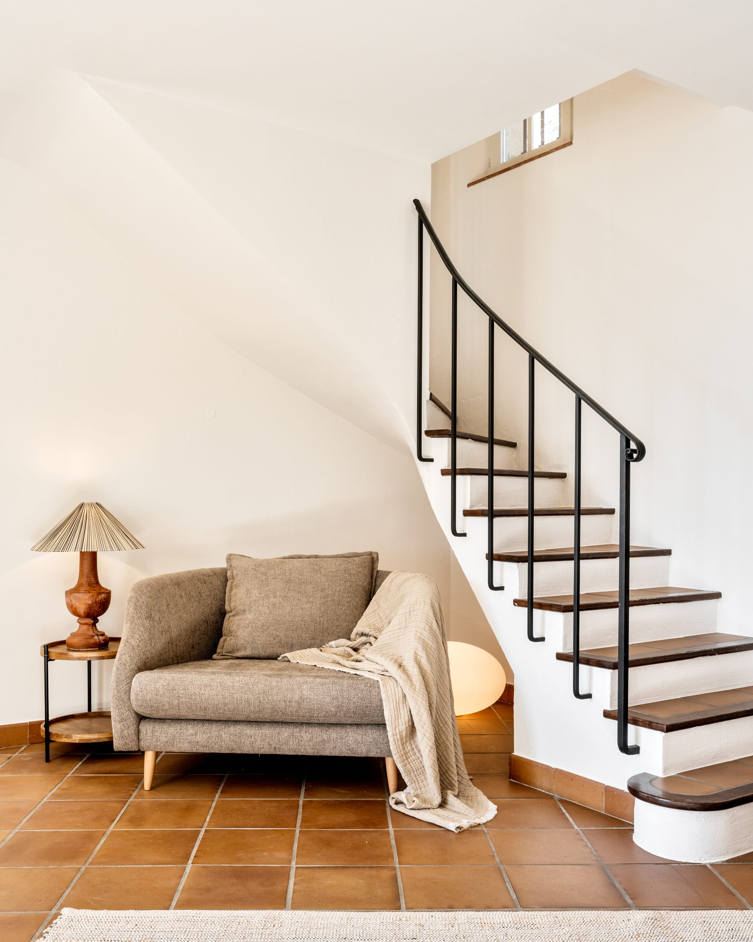 Cosy reading corner with a linen loveseat, ceramic table lamp, and a curved staircase