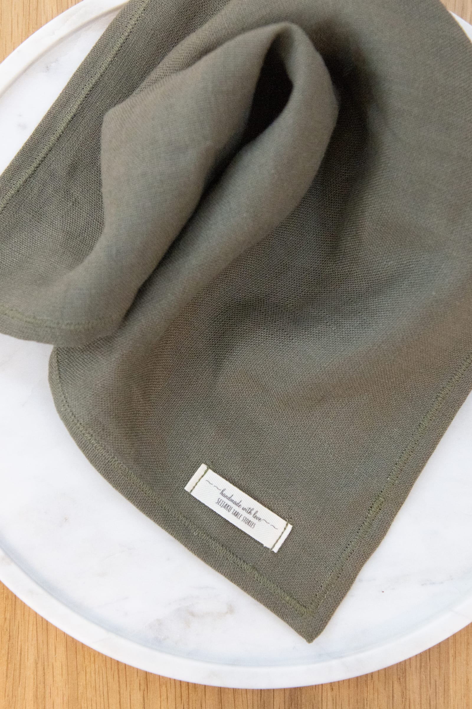 Folded earthy CLAY-green linen napkin with hand-finished corners, on a ceramic plate