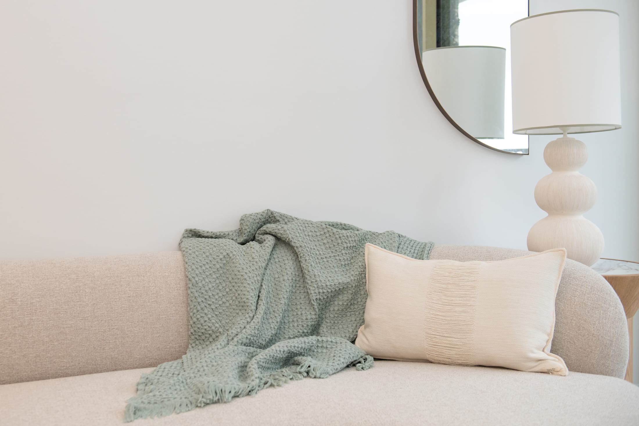 The cushion styled on a sofa with a sage-green waffle throw, beneath a round mirror and ceramic table lamp